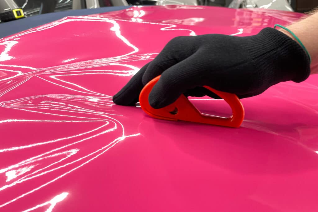 Ein Spezialist für die Umhüllung eines Autos mit rosafarbener Vinylfolie während der Arbeit. Car-Wrapping-Spezialisten bedecken das Auto mit Vinylfolie oder -folie. Autoverpackung aus nächster Nähe. Selektiver Fokus.